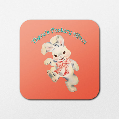 The Fuckery Afoot Coaster by Blue Dog Posters features a vintage-style illustration of a smiling white rabbit with a pink ribbon. The playful text "That's Fuckery Afoot" appears above, set against a soft coral background.