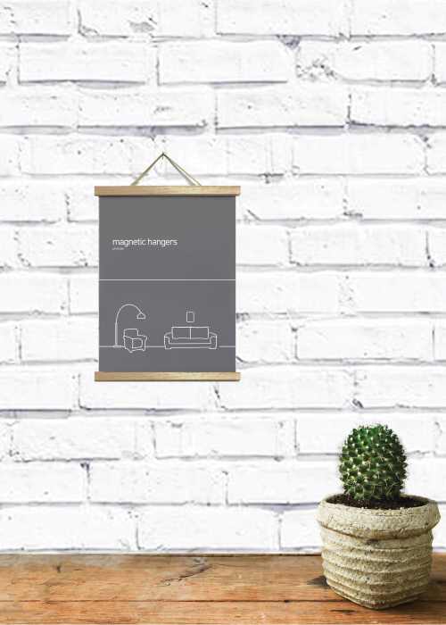 A decorative poster with a minimalist design hangs on a white brick wall. Held by the Blue Dog Posters Magnetic Timber Hanger 15cm Natural, the poster features a simple line drawing of furniture and the text "magnetic hangers." Below, on a wooden surface, a small potted cactus adds a touch of greenery.