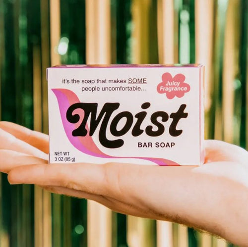 A person's hand is holding a box of "Moist Triple Milled Boxed Bar Soap" with "Juicy Fragrance," crafted by Blue Dog Posters. The Vermont-made soap features a box design with pink and brown text. The tagline reads, "it's the soap that makes SOME people uncomfortable..." The background has blurred vertical green and light brown stripes.