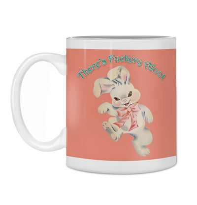 The Hustle and Co Mug - Fuckery Afoot from Blue Dog Posters, part of our Homewares Collection, features a vintage-style illustration of a rabbit with a pink bow. Above the rabbit, playful turquoise text reads, "There's Fuckery Afoot," on a muted reddish-brown background that enhances the ceramic finish.