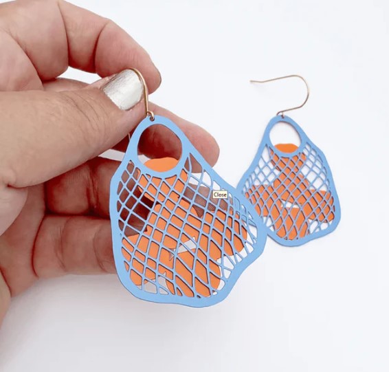 A hand holds Blue Dog Posters’ String Bag Earrings in Blue with Oranges, featuring lightweight blue mesh over orange shapes and hypoallergenic hooks, set against a plain white background.