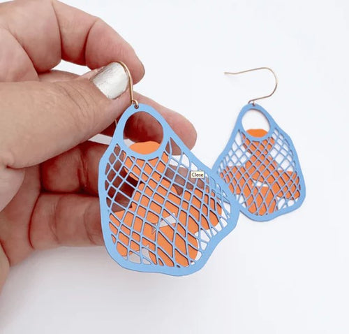 A hand holds Blue Dog Posters’ String Bag Earrings in Blue with Oranges, featuring lightweight blue mesh over orange shapes and hypoallergenic hooks, set against a plain white background.