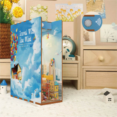 The Travel With the Wind Book Nook by Blue Dog Posters, featuring a city, houses, hot air balloons, and vibrant colors, sits on a bedside table in a cozy bedroom decorated with mini houses and an alarm clock.