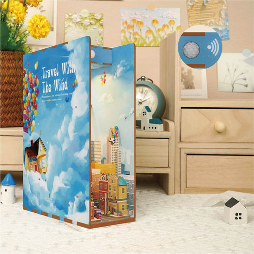 The Travel With the Wind Book Nook by Blue Dog Posters, featuring a city, houses, hot air balloons, and vibrant colors, sits on a bedside table in a cozy bedroom decorated with mini houses and an alarm clock.