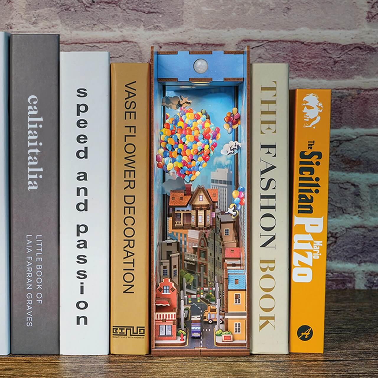 A row of books on a shelf features the Blue Dog Posters "Travel With the Wind Book Nook Tonecheer" diorama—colorful houses and balloons inspired by "Up"—nestled among the books, with a brick wall in the background.
