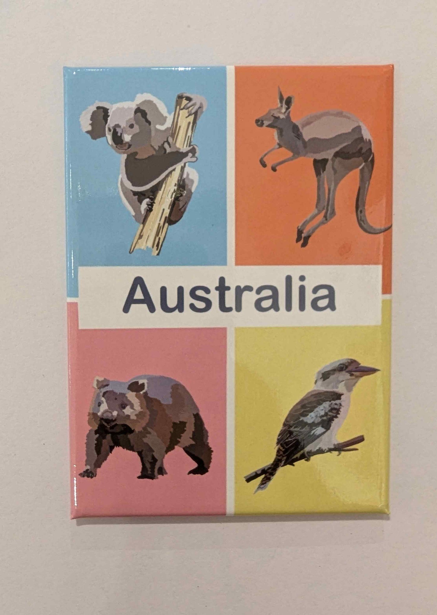 Brighten your fridge with the Travel Magnet - Australia Animals by Blue Dog Posters, featuring a koala, kangaroo, wombat, and kookaburra in four sections with "Australia" at the center—a perfect travel souvenir.