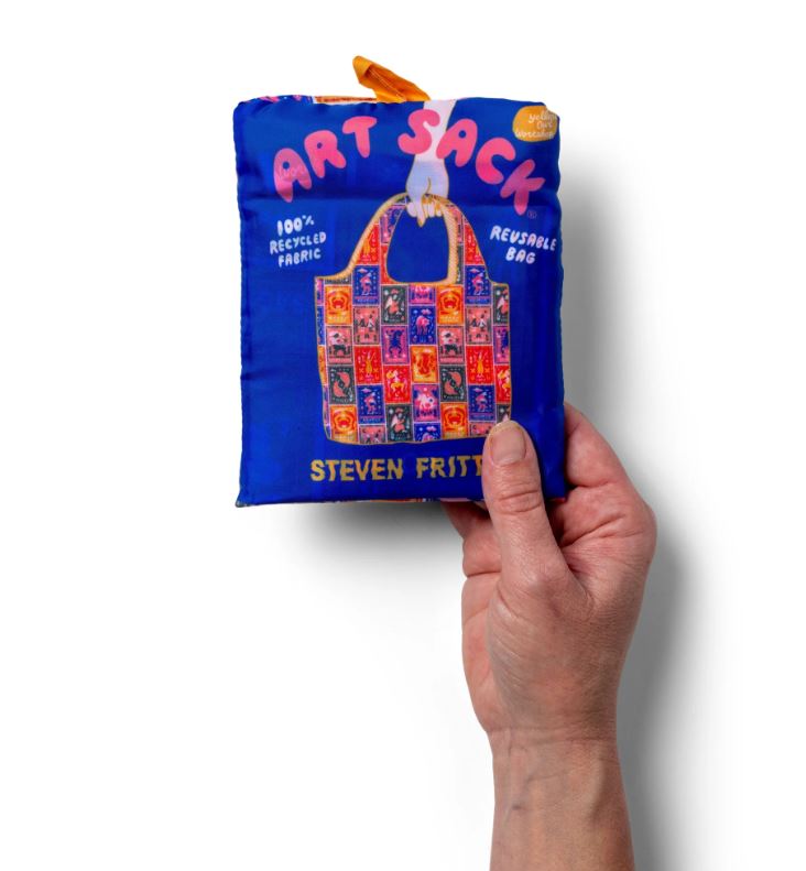 A hand holds a folded blue Zodiac Art Sack by Blue Dog Posters, made from 100% recycled rPET. The eco-friendly bag features vibrant artwork and is labeled "REUSABLE BAG," making it a perfect astrology-themed gift.