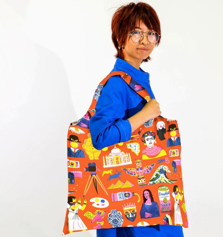 A person with short, reddish hair and glasses stands against a plain white background. They are wearing a bright blue outfit and holding a large, colorful Art History Art Sack tote bag over their shoulder. The machine washable bag from Blue Dog Posters is made from 100% recycled rPet and features vibrant designs with various cultural symbols and icons.