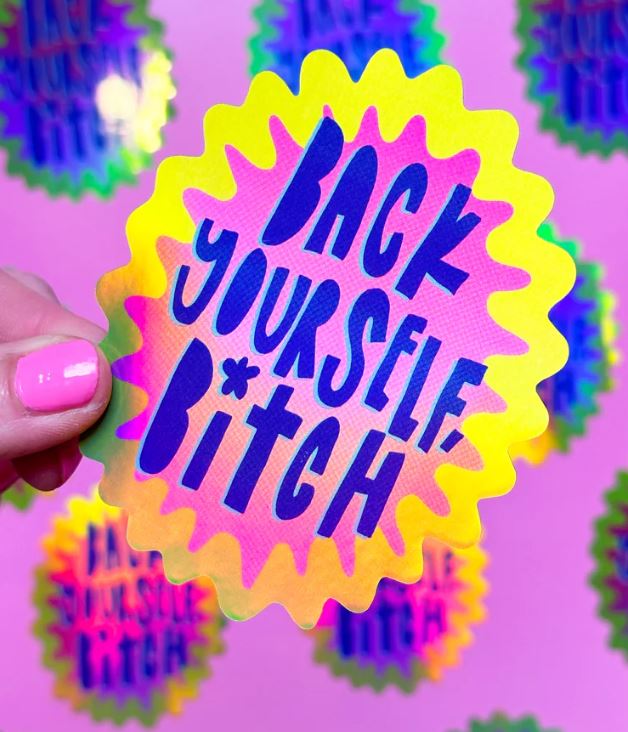A hand with pink nail polish holds a Blue Dog Posters Back Yourself Bitch Holographic Sticker. The vibrant pink background is scattered with similar stickers designed by Bea Bellingham.