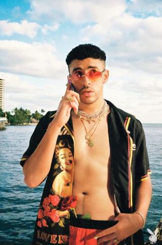 A man in sunglasses, gold chains, and an open graphic shirt stands by the ocean holding a phone—ideal as a Bad Bunny music poster from MUS 296, featuring palm trees and city buildings under partly cloudy skies.