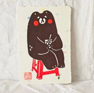 Bear Red Chair Small Lino Print by Misu-Juju from Blue Dog Posters features a cute black bear with red cheeks on a red chair, sipping a drink. This charming design sits on a white background with soft colorful spots for a lino print feel.