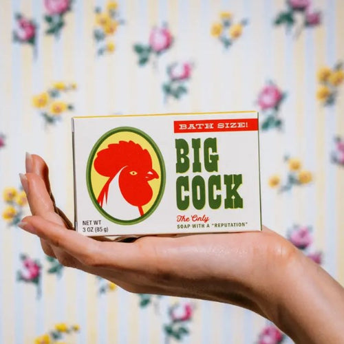 A person holds a bar of Big Cock Triple Milled Boxed Bar Soap with retro packaging labeled "Bath Size! Big Cock" in bold green text and an image of a rooster. The background features floral wallpaper with yellow and white stripes and pink flowers, embodying the playful spirit of Blue Dog Posters.
