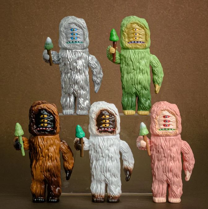 The Blue Dog Posters BigWoot Figure Blind Box features five colorful, furry, three-eyed monster figurines in suits of gray, green, brown, white, or pink—each holding a tree-shaped item in a whimsical folklore-inspired display.