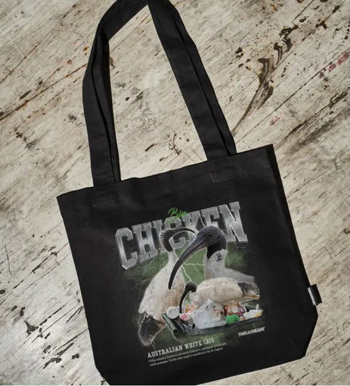 The Vintage Bin Chicken Tote Bag Black by Blue Dog Posters is a black cotton canvas tote featuring an Australian white ibis eating from a bin and "Bin Chicken" text. Ethically sourced and displayed on a textured, weathered floor.