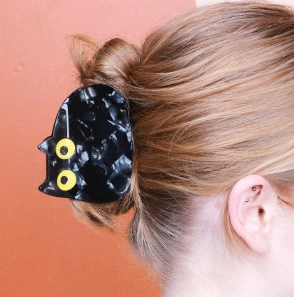 A person with light brown hair in a bun, secured by the Void Black Cat Hair Claw from Blue Dog Posters, stands against a reddish-brown wall—a scene echoing the quirky charm of Bea Bellingham’s collection.