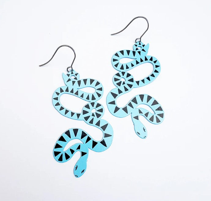 The Snake Earrings in Blue Black by Denz from Blue Dog Posters feature coiled snakes in light blue with black geometric patterns, detailed eyes, and open mouths—unique wearable art with hooks for easy wearing.