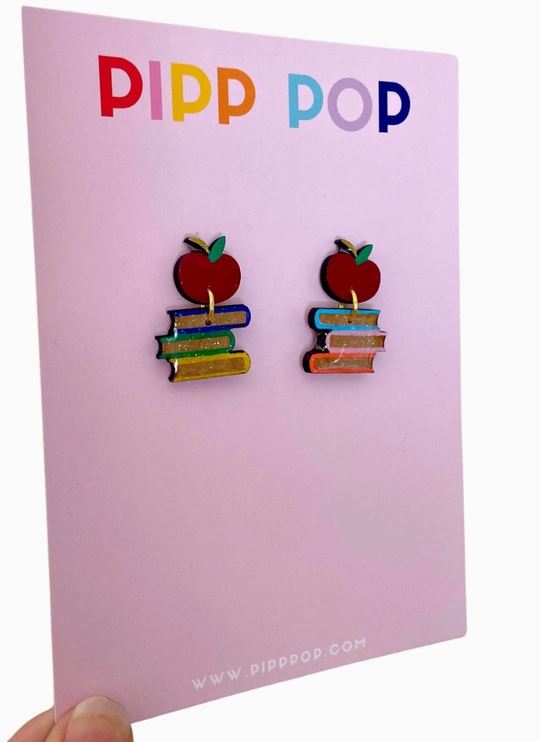 A hand holds a pink "PIPP POP" card displaying Blue Dog Posters' Book Dangle Earrings—handmade earrings shaped like colorful stacks of books with a red apple on top, making them perfect teacher gifts or book lover accessories.