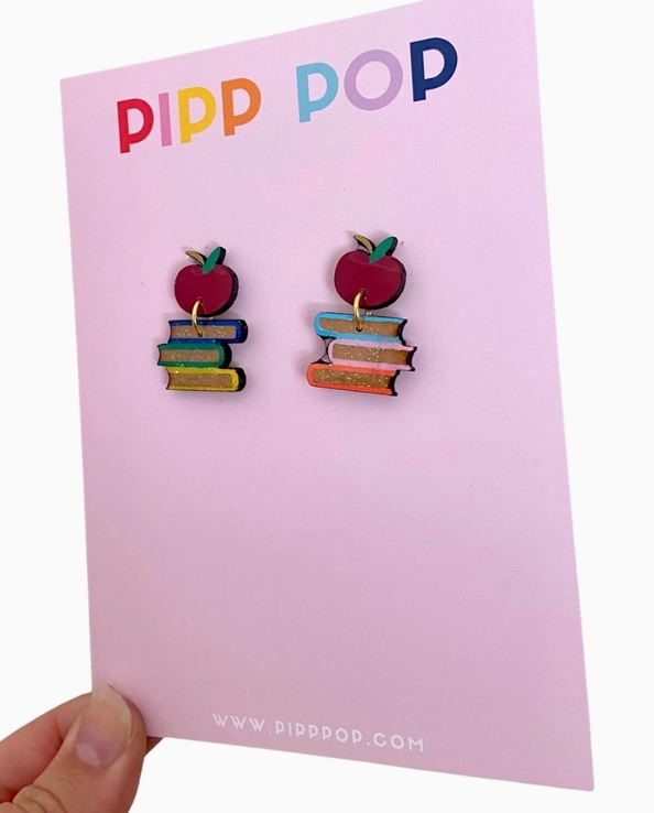 A hand holds a card displaying Book Dangle Earrings shaped like red apples on colorful books, with the "Blue Dog Posters" logo at the top.