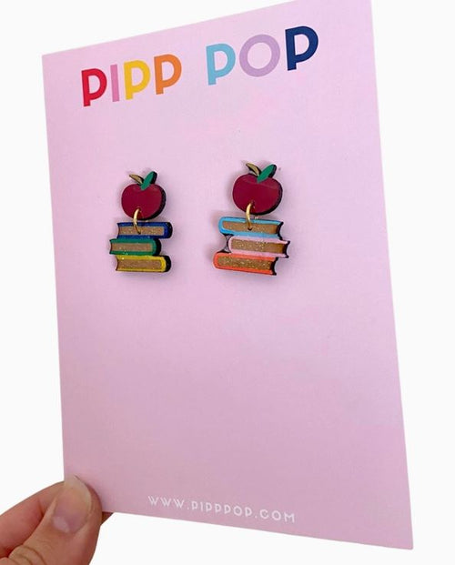 A hand holds a card displaying Book Dangle Earrings shaped like red apples on colorful books, with the "Blue Dog Posters" logo at the top.