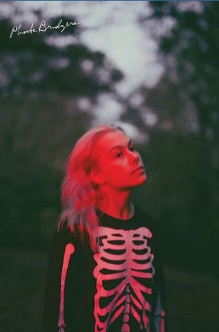 A person with light hair in a black skeleton shirt stands outside at dusk in red light, gazing upward. The scene resembles the MUS 303 Phoebe Bridgers - Punisher poster printed on premium gloss paper, with blurry trees and clouds behind.