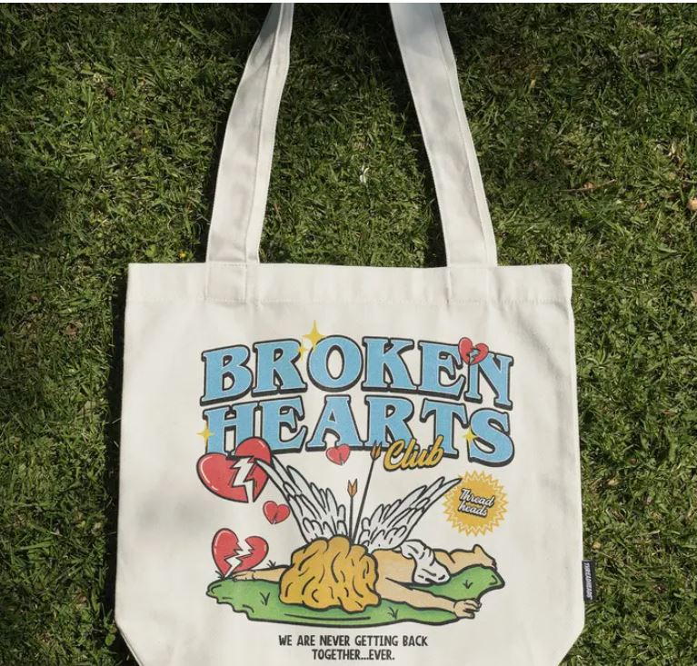 A white cotton canvas tote by Blue Dog Posters lies on grass, displaying colorful cartoon art of a broken heart and brain with "BROKEN HEARTS Club" and "WE ARE NEVER GETTING BACK TOGETHER. EVER." Product: Broken Hearts Club Tote Bag.