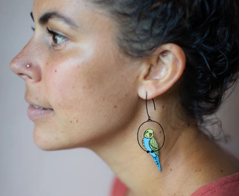 A woman with curly dark hair and a nose stud wears large Budgie Black Blue Yellow Denz Earrings by Blue Dog Posters, made from recycled stainless steel. Shown in profile, she gazes left with a neutral expression.