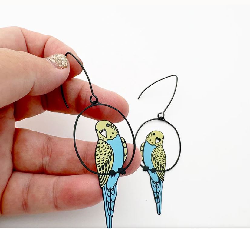 A hand displays the Budgie Black Blue Yellow Denz Earring by Blue Dog Posters inside a black hoop, crafted from recycled stainless steel, with its lightweight match resting on a white background.