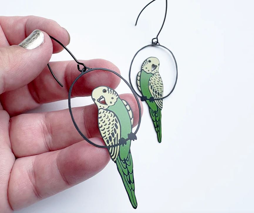 A hand holds a Budgie Black Green Yellow Denz Earring by Blue Dog Posters, featuring a green and yellow parakeet design; a matching lightweight earring made from recycled stainless steel hangs nearby against a white background.