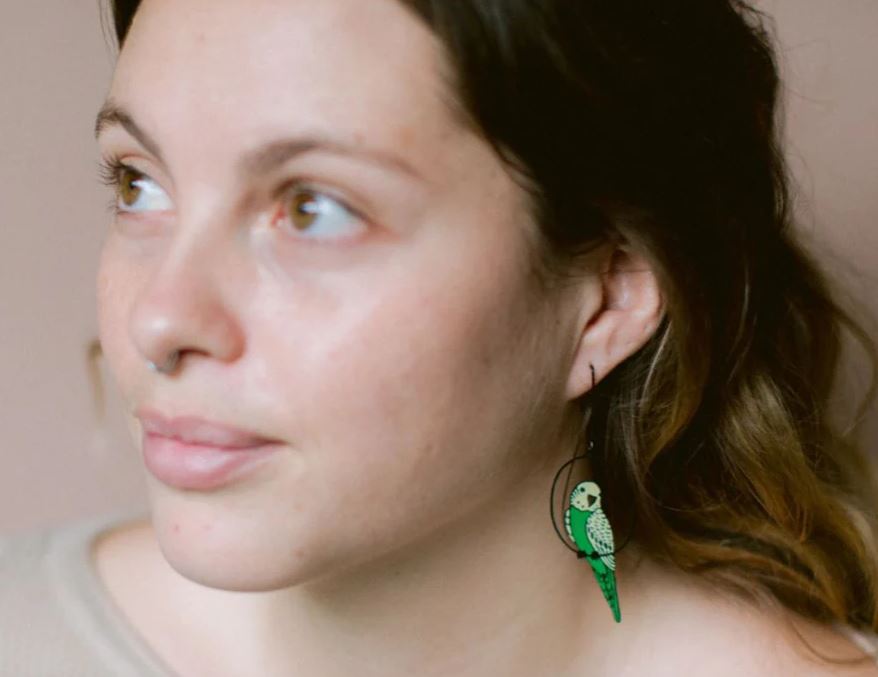 A person with wavy brown hair gazes sideways, wearing Budgie Black Green Yellow Denz Earrings by Blue Dog Posters, crafted from recycled stainless steel. A calm, thoughtful expression and a light neutral background complete the scene.