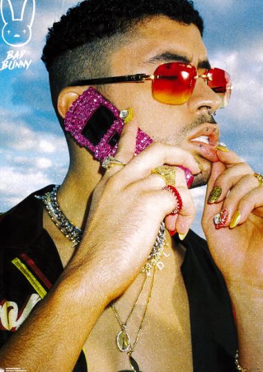 A person with short curly hair and red-tinted sunglasses holds a pink rhinestone flip phone, striking an A3 music poster pose for the Bad Bunny by Blue Dog Posters against a partly cloudy sky.