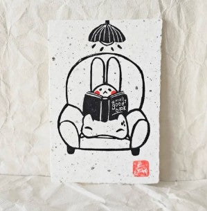 Bunny Book Chair Small Lino Print by Misu-Juju for Blue Dog Posters features a minimalist rabbit reading in an armchair under a tiny umbrella, with a textured white paper background reminiscent of hand-pressed lino prints.