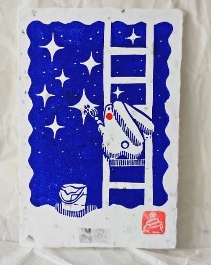 The Bunny Painting Stars Small Lino Print by Misu-Juju from Blue Dog Posters shows a white rabbit climbing a ladder and painting stars against a blue night sky, with a paint can below. This unique art print features an original blue and white design.
