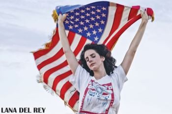 A woman stands outdoors, eyes closed, holding an American flag overhead and wearing the Lana Del Rey - Flag and Buttwiser Shirt by MUS 018. The sky is clear behind her. "LANA DEL REY" features in the bottom left—perfect for music fans!.