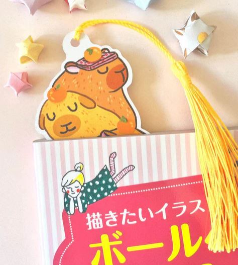 The Bookmark Capybara Stack by Jess Chen from Blue Dog Posters—a die-cut bookmark with stacked capybaras and a yellow tassel—rests in a pink-striped book, surrounded by colorful origami stars and a girl illustration on the cover.
