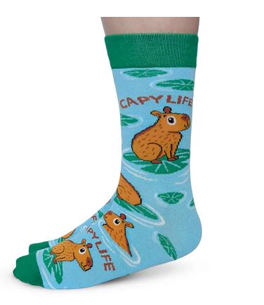 A person is wearing Blue Dog Posters Capy Life Socks—women's cotton socks with a green cuff, reinforced toe, and cartoon capybaras on lily pads with "CAPY LIFE" text on a light blue background.