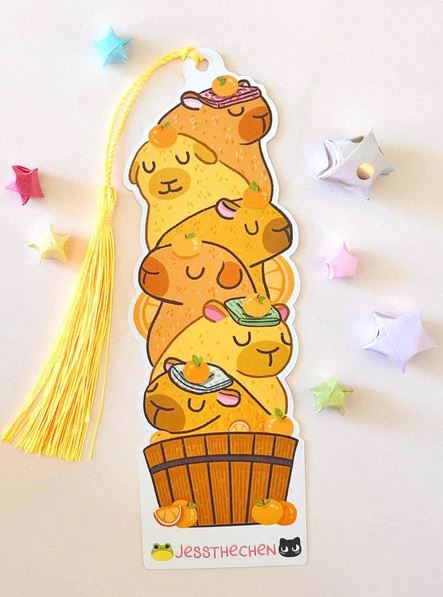 The Bookmark Capybara Stack by Jess Chen from Blue Dog Posters features cute cartoon capybaras in a wooden tub with fruit hats, die-cut on premium cardstock with pastel stars and a yellow tassel.