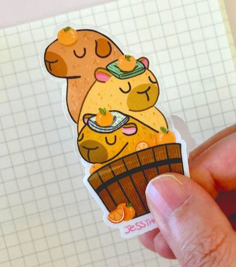 A hand holds the Blue Dog Posters Capybara Stack Vinyl Single Sticker by Jess Chen, featuring cute capybaras stacked in a wooden tub with oranges on their heads. The waterproof vinyl sticker is set against graph paper.