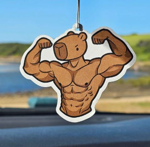 The Blue Dog Posters Muscle Capybara Orange Air Freshener, shaped like a flexing capybara, hangs in a car against a blurred outdoor view—ideal for fans of quirky cartoon designs and unique air fresheners.