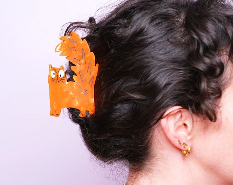 A person with dark curly hair wears an orange Azz Hole Angry Cat Hair Claw by Blue Dog Posters. The photo shows the side of their head and gold hoop earrings against a light pink background.