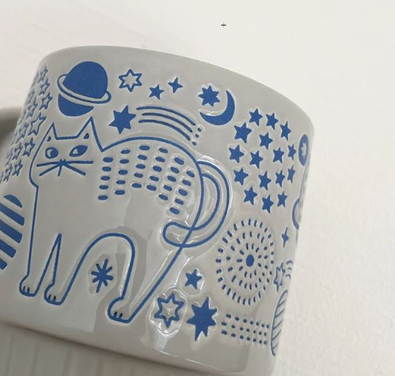 The Cat Flower Garden Stone & Blue Japanese Mug by Blue Dog Posters features a ceramic design with blue cat and celestial motifs on a light background, inspired by Japanese zakka style.