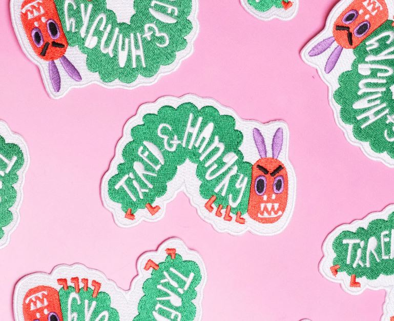 Blue Dog Posters' Tired and Hungry Catapillar Patch features a green caterpillar with a red face, bunny ears, and "Tired & Hungry" text—an embroidered patch inspired by Bea Bellingham's playful style, shown on a pink background.