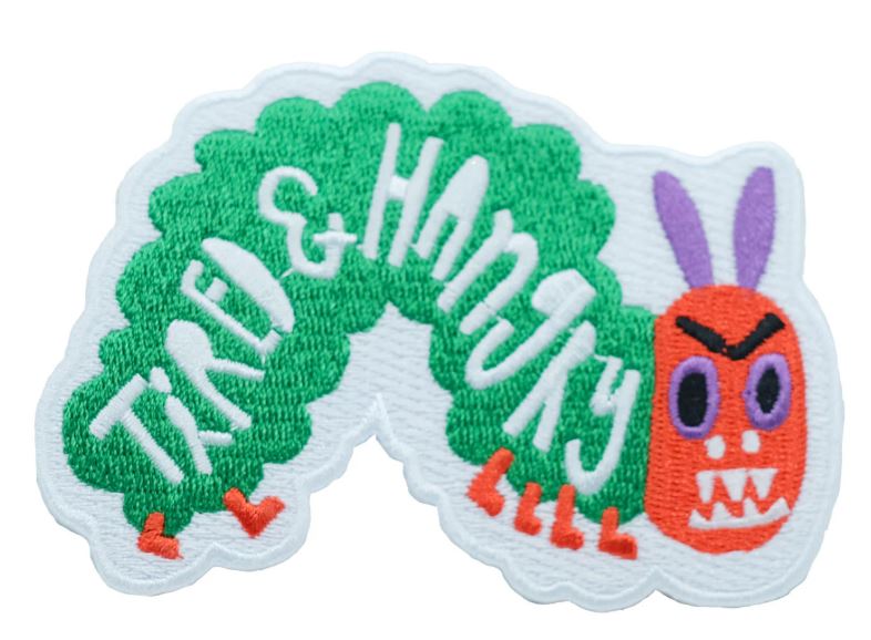The Tired and Hungry Caterpillar Patch by Blue Dog Posters features a green caterpillar with an orange face, purple ears, and a grumpy look. "TIRED & HANGRY" is embroidered in white—a playful touch inspired by Bea Bellingham’s quirky style.