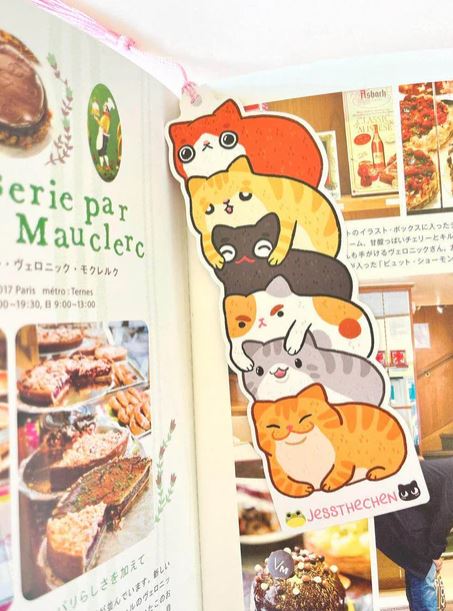 The Bookmark Cat Stack by Jess Chen from Blue Dog Posters, shaped like five colorful cartoon cats, is placed in an open food magazine to mark a page with photos of pastries and French text.