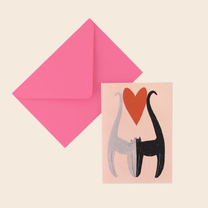 Introducing the Cats Love Card by Blue Dog Posters: This charming greeting card depicts two cats, one gray and one black, with their tails intertwining to form a heart shape. Above the duo is a bold red heart. Designed by Little Black Cat Illustrated in Manchester, UK, this card comes with a vibrant pink envelope.