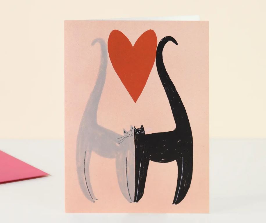 The Cats Love Card by Blue Dog Posters showcases two stylized cats, one grey and one black, standing side by side with their tails forming an outline of a heart. Above them is a large red heart against a light peach background. A closed pink envelope is also partially visible. From Little Black Cat Illustrated in Manchester, UK.