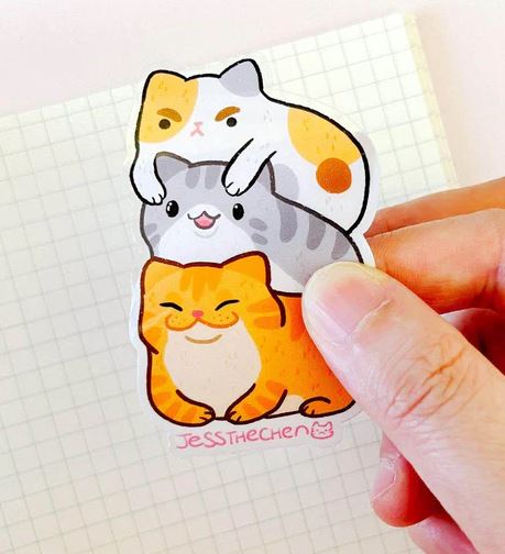 A hand displays the Cat Stack Vinyl Single Sticker by Jess Chen from Blue Dog Posters, featuring three cartoon cats stacked together on grid paper. This waterproof vinyl sticker is ideal for any cat lover!.