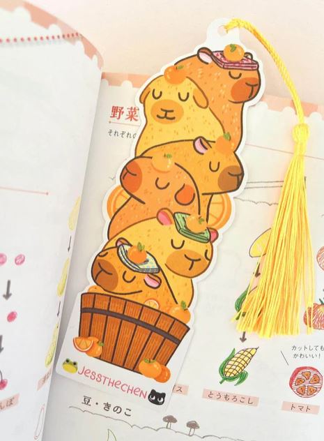 The Bookmark Capybara Stack by Jess Chen from Blue Dog Posters features four cartoon capybaras stacked in a wooden tub, each holding food, with a die-cut design and yellow tassel—perfect for any illustrated book.
