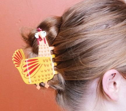 A person with light brown hair wears a bun secured by the Screaming Chicken Hair Claw from Blue Dog Posters. The bright yellow clip, made from an acetate alternative, stands out against a solid reddish background.