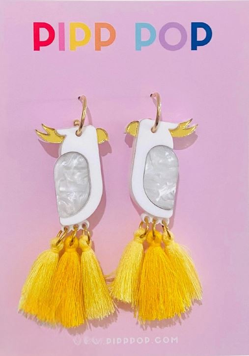 White Cockatoo Dangles by Blue Dog Posters: playful, hypoallergenic earrings with white bodies, gold wings, and yellow tassels, shown on a pink card topped with "PIPP POP" in colorful letters.