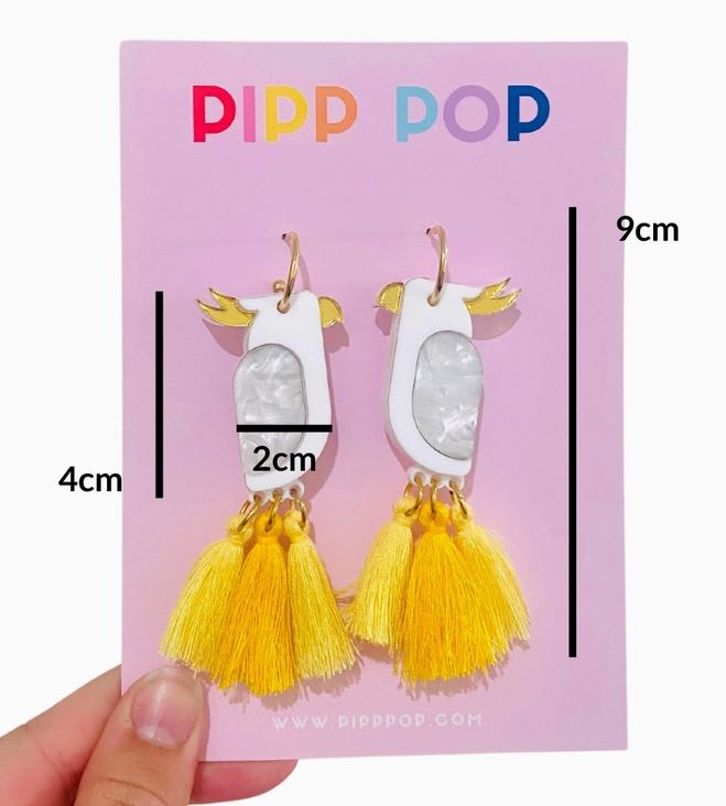 A hand holds a PIPP POP card with Blue Dog Posters’ White Cockatoo Dangles—hypoallergenic earrings featuring white bodies, gold beaks, yellow tassels; measuring 9cm long, 4cm wide, and 2cm at the body.